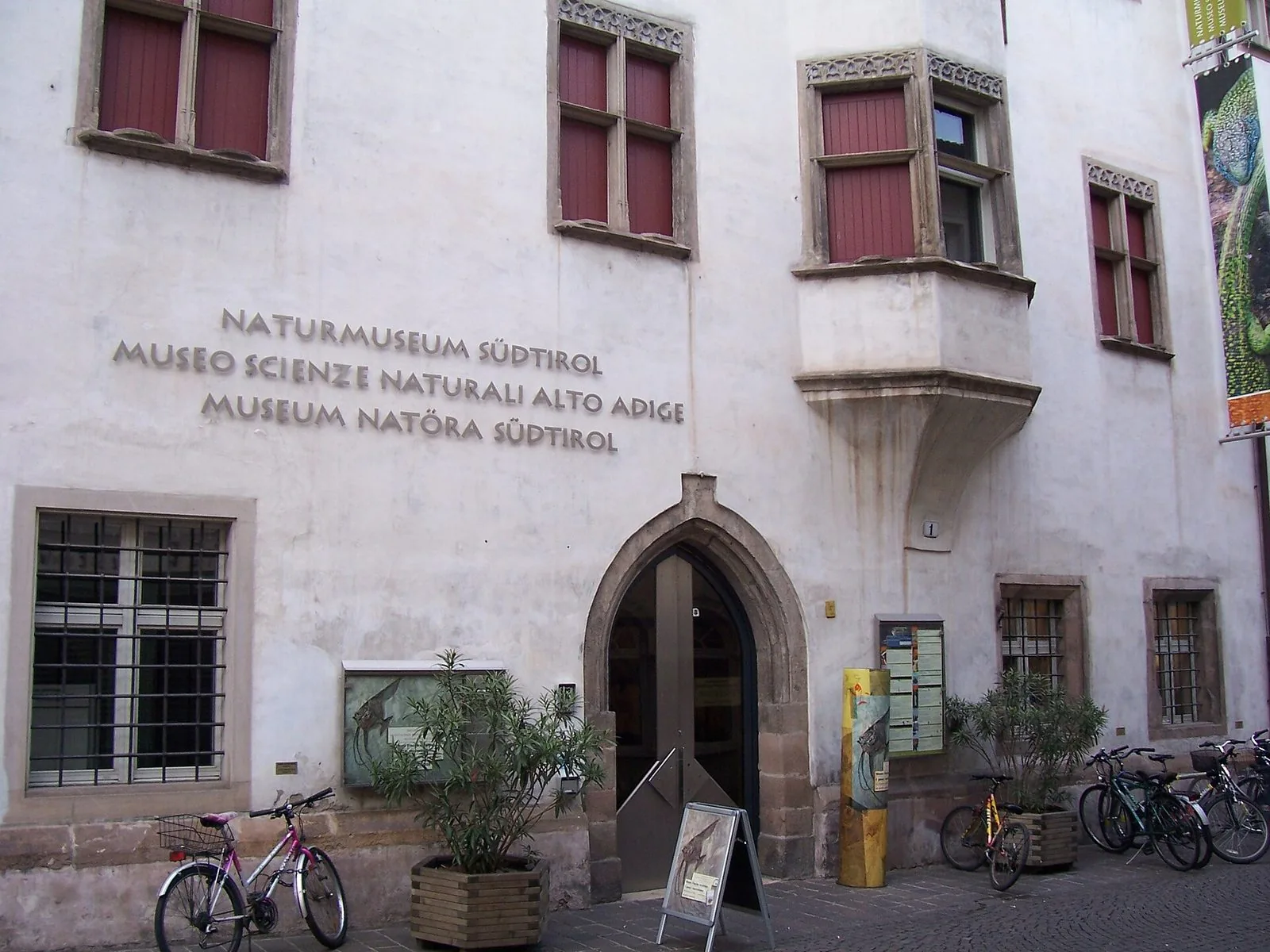 La facciata bianca del Museo di Scienze Naturali dell'Alto Adige a Bolzano, con finestre in pietra dalle imposte rosse, un erker sporgente, l'insegna trilingue in rilievo, il portale gotico d'ingresso, fioriere e biciclette parcheggiate davanti