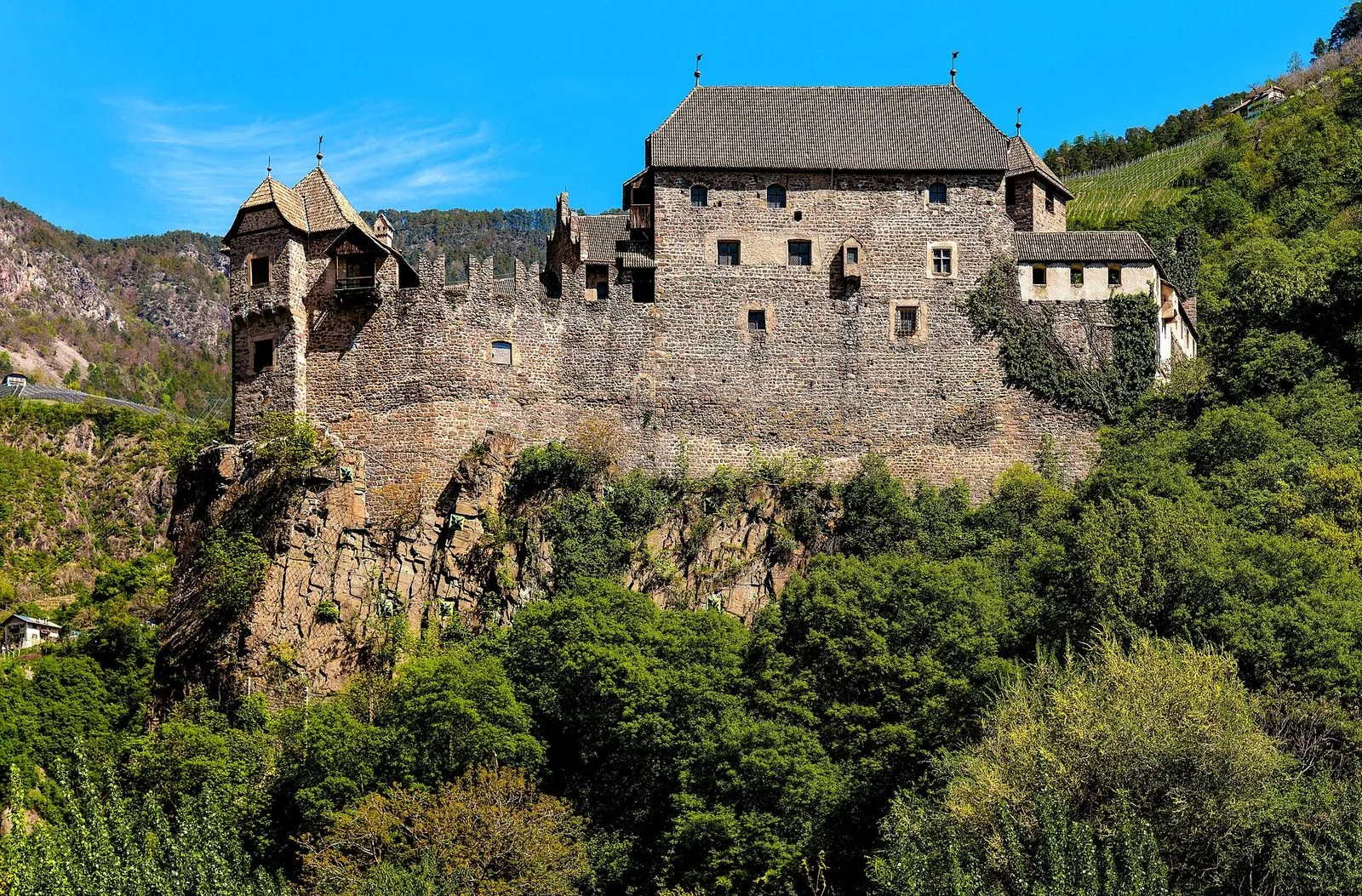Castel Roncolo visto dal basso: le mura merlate in pietra, la torretta angolare e il grande edificio dal tetto scuro poggiano su uno sperone di roccia, circondati da boschi verdi e vigneti sotto un cielo azzurro