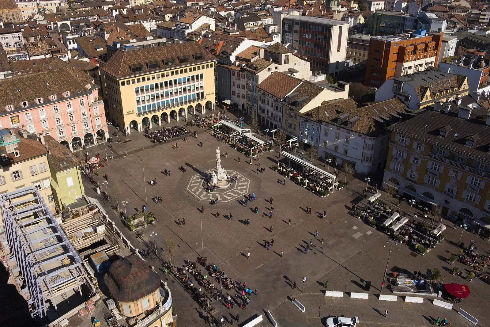 Piazza Walther a Bolzano vista dall'alto: al centro il monumento a Walther von der Vogelweide su un disegno ottagonale nella pavimentazione, dehors coperti e passanti attorno, palazzi storici dai tetti in tegole e l'edificio porticato della Cassa di Risparmio