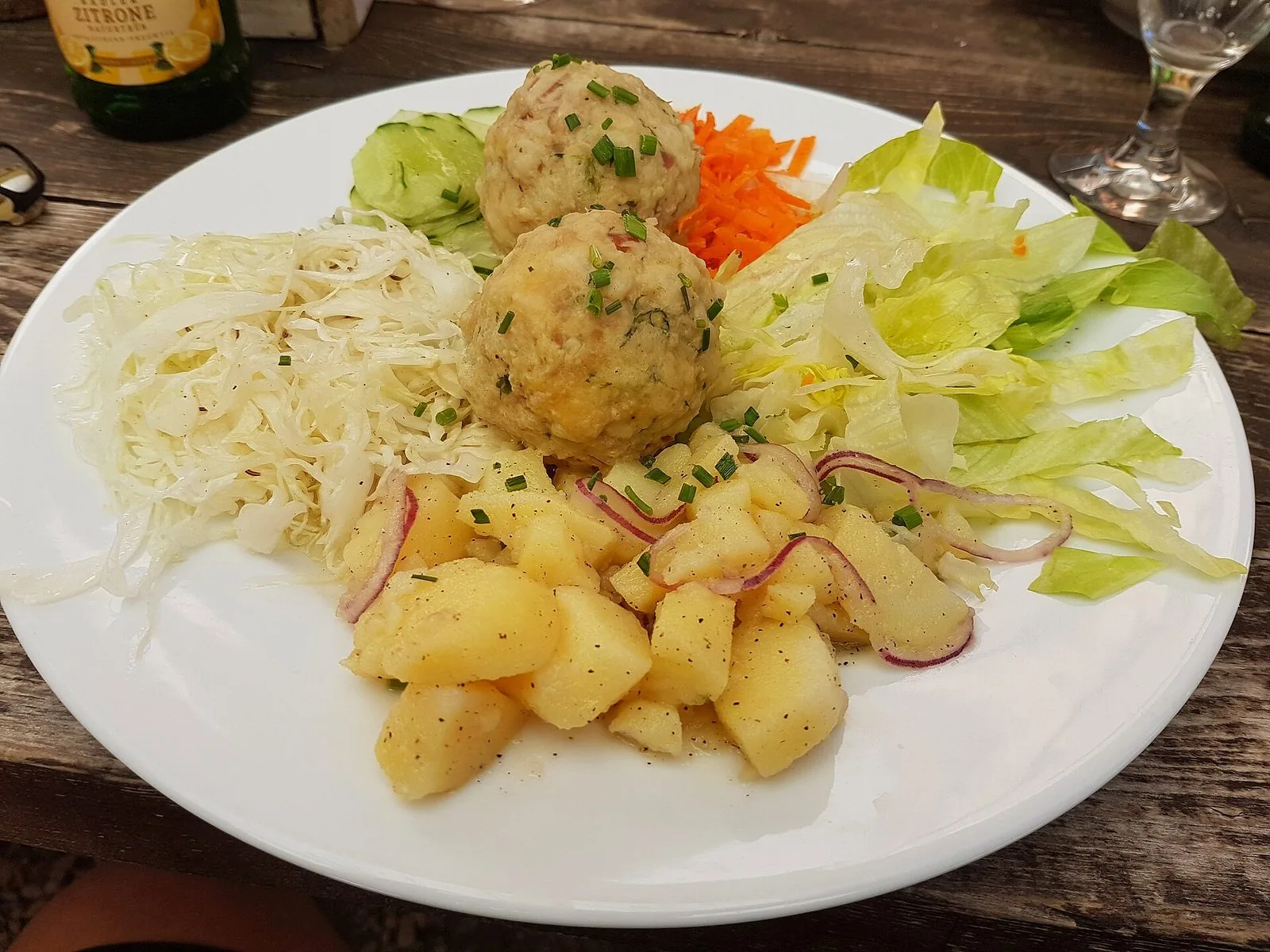 Un piatto di canederli allo speck serviti con insalata mista: cavolo cappuccio, lattuga, carote e patate condite con cipolla rossa, su un tavolo di legno