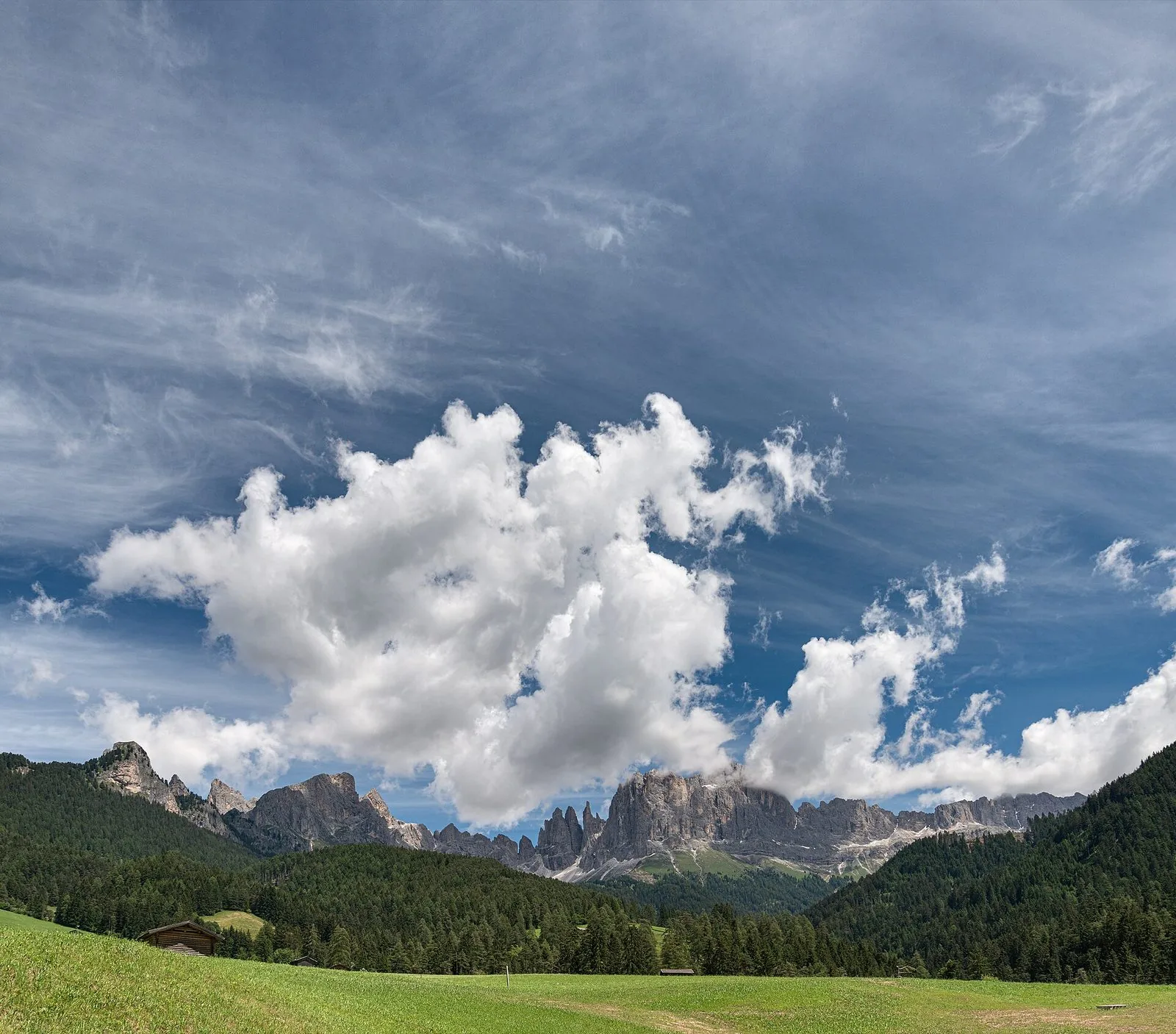 Il gruppo del Catinaccio visto dalla Val di Tires: pareti e guglie di roccia chiara, fra cui le sottili Torri del Vajolet, sopra boschi di conifere e un prato verde con un fienile in legno, sotto un ampio cielo blu con grandi nuvole bianche