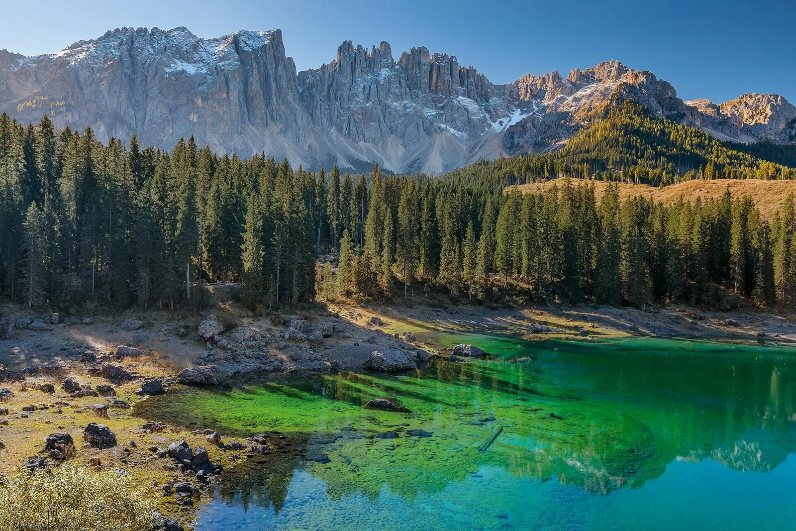 Il Lago di Carezza con l'acqua limpida verde smeraldo e turchese, la riva sassosa, un fitto bosco di abeti e sullo sfondo le pareti e le guglie del Latemar spruzzate di neve sotto un cielo azzurro
